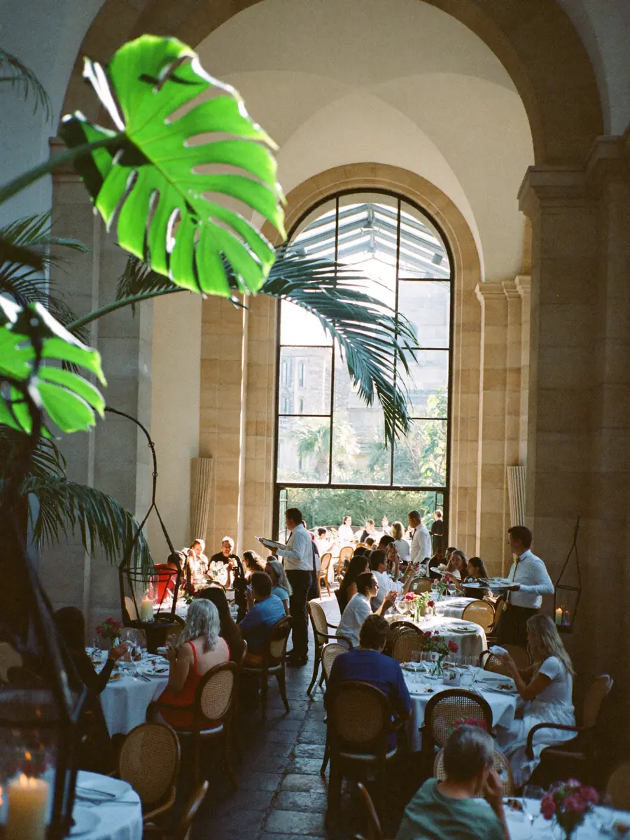 A sunlit luxury dining room seen from the entrance archway, every table taken and two servers working the floor